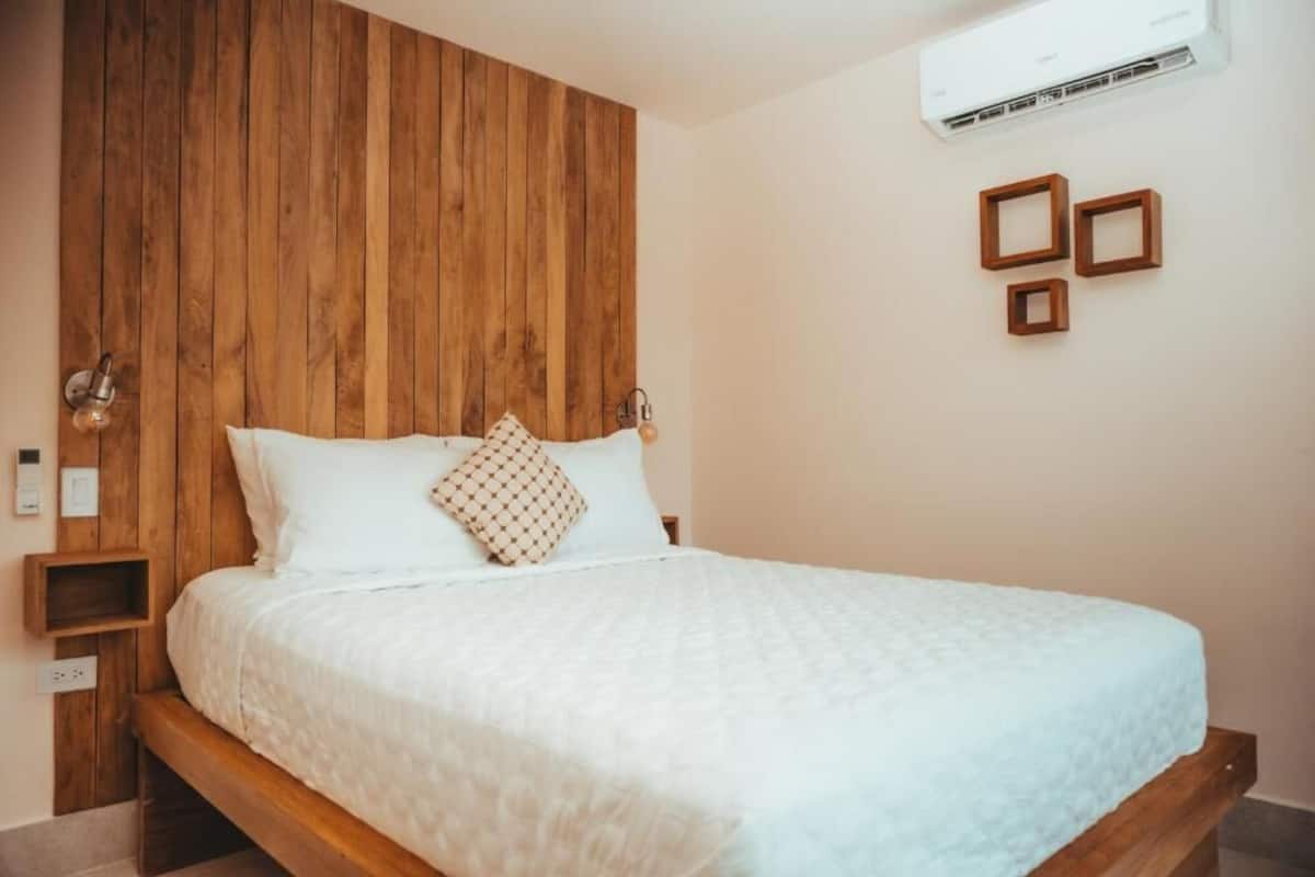 Une chambre à coucher avec une tête de lit en bois, un lit avec une literie blanche, une petite table de chevet et un climatiseur fixé au mur.