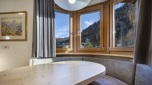 A room with a round table, a cushioned bench, and large windows offering a mountain view. - 1012A Apartments Monica (Santa Cristina Valgardena)