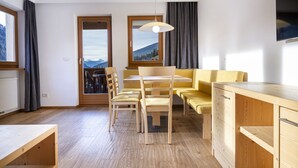 A modern kitchen with wooden cabinets, a dining table with chairs, and a view of mountains through the windows. - 1012A Apartments Monica (Santa Cristina Valgardena)