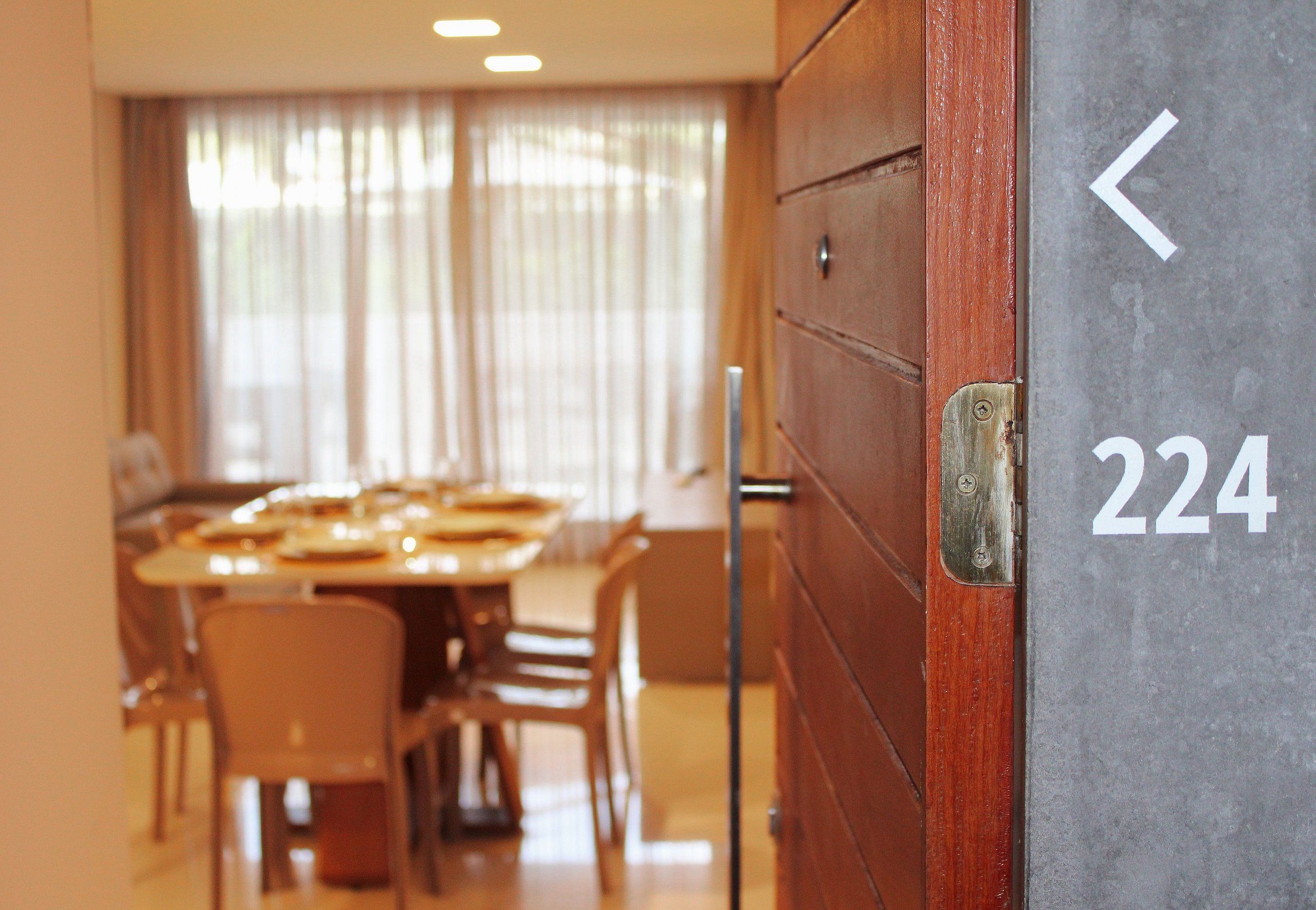 L’entrée d’une chambre d’hôtel, avec une porte en bois, le numéro « 224 » affiché sur le mur, et un coin repas avec des tables et des chaises à l’arrière-plan.