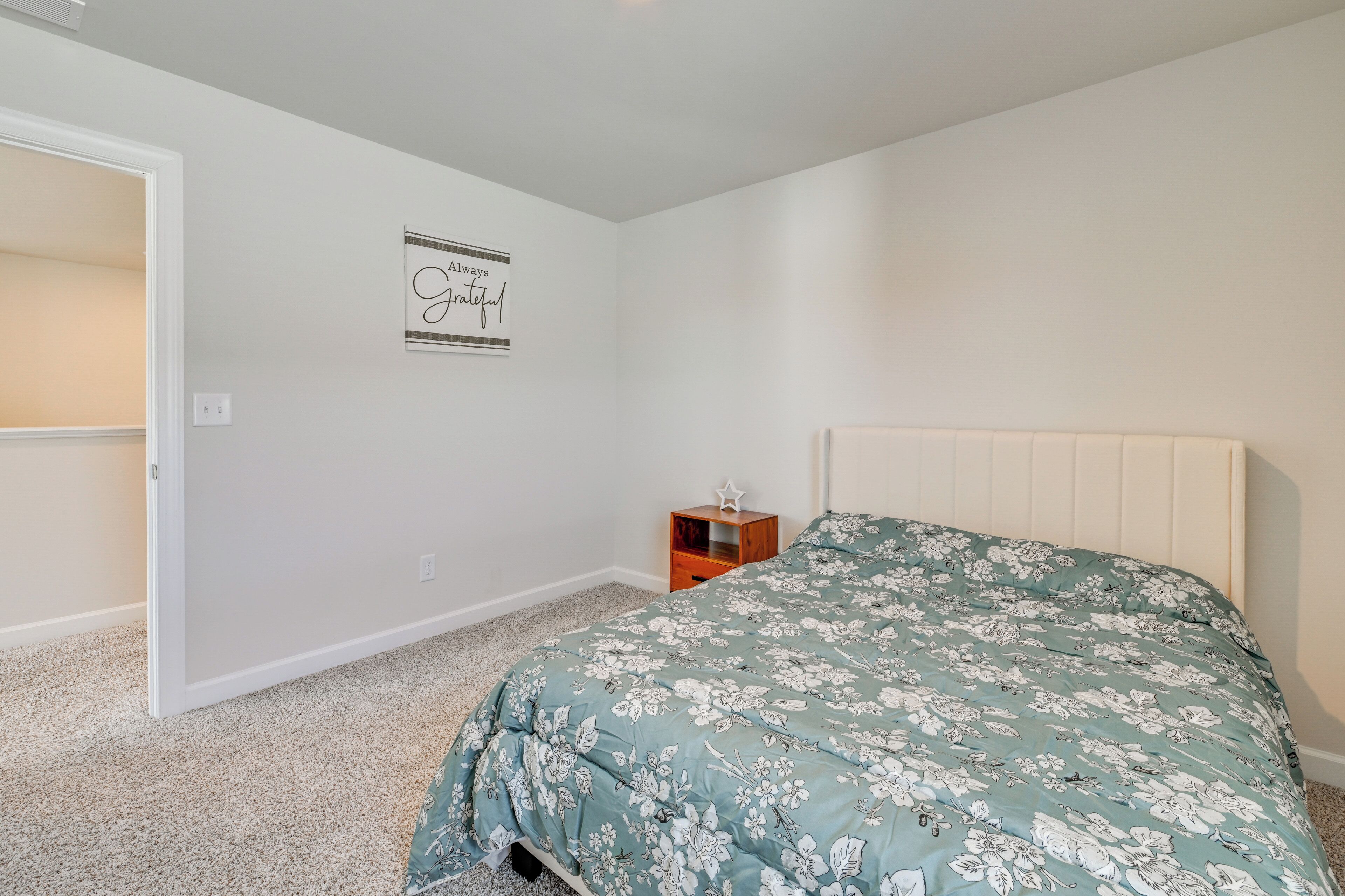 A bedroom with a floral bedspread, a white headboard, a small wooden nightstand, and a wall with a motivational quote.