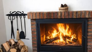 A fireplace with a brick surround and a black frame, a basket of firewood, and a hanging rack with tools.