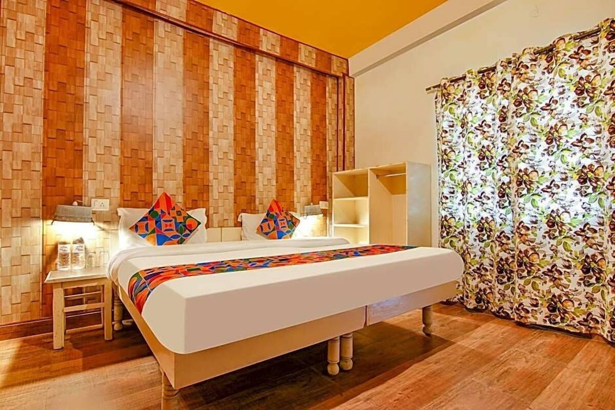 A hotel room with a wooden wall, a bed with colorful bedding, a nightstand, a lamp, and a floral curtain.