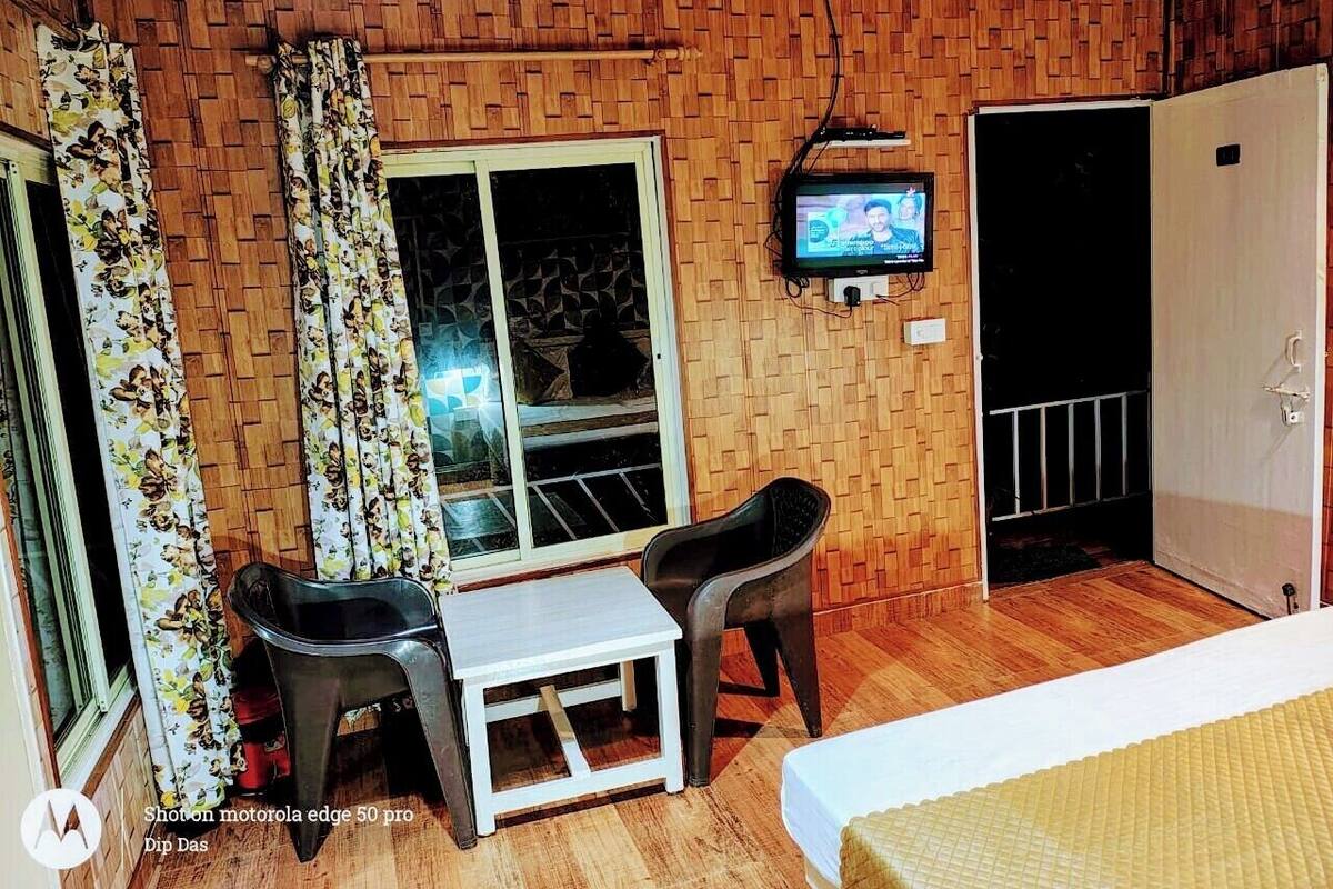 A room with a wooden wall, a television mounted on the wall, a bed, a table, two chairs, and a window with curtains.