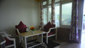 A room with a wooden table and two chairs, a small table with a vase of flowers, and a window with floral curtains. - The Perfect Hideout By Himalayan Grove (Dehradun)