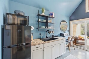 A modern kitchen with a built-in microwave, a stainless steel refrigerator, a wooden countertop, and a black sink. There is a shelf with decorative items and a potted plant.