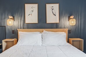 Ein Bett mit weißer Bettwäsche, zwei Wandlampen und zwei Bilder von Vögeln in Rahmen an einer dunklen Wand.