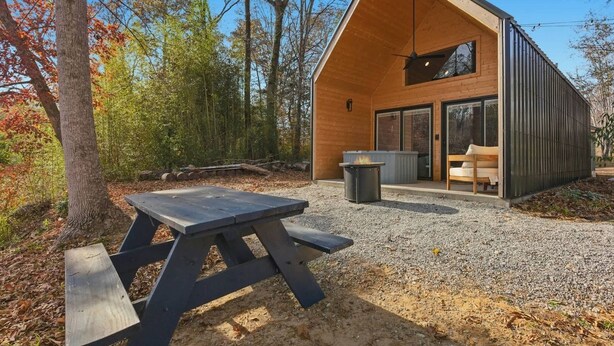 A modern cabin with a picnic table, a fire pit, and a large window overlooking a forest.