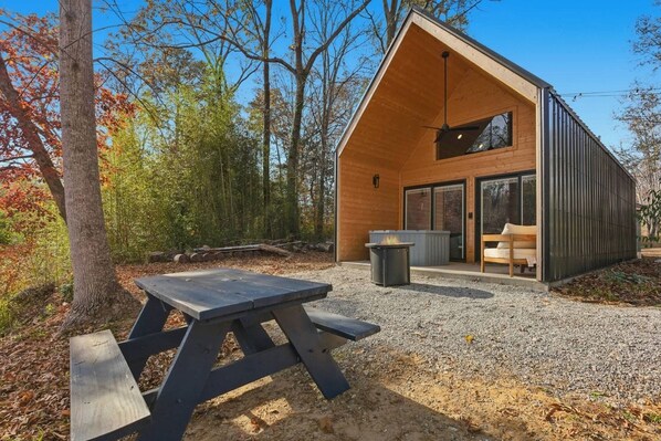 A modern cabin with a picnic table, a fire pit, and a large window overlooking a forest.