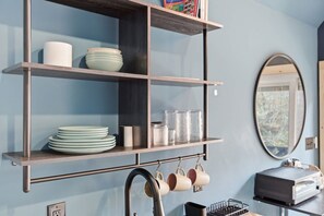 A kitchen with a shelf holding plates and bowls, a hanging faucet, and a mirror on the wall.