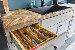 A kitchen with a herringbone wood countertop, a black sink, and a drawer with various utensils.