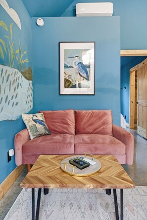 A room with a pink sofa, a framed bird painting, and a wooden coffee table with a remote control. - The Blue Heron - Riverfront Retreat w/ Hot Tub (Easley)