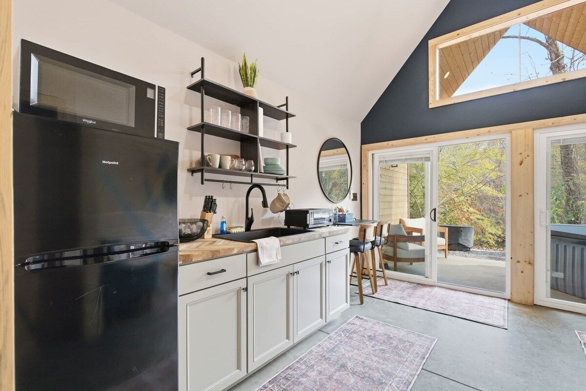 A modern kitchen with a black refrigerator, white cabinets, and a wooden countertop. There is a built-in microwave above the stove, and a shelf with glassware and a plant.