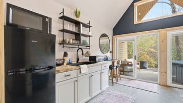 A modern kitchen with a black refrigerator, white cabinets, and a wooden countertop. There is a built-in microwave above the stove, and a shelf with glassware and a plant.