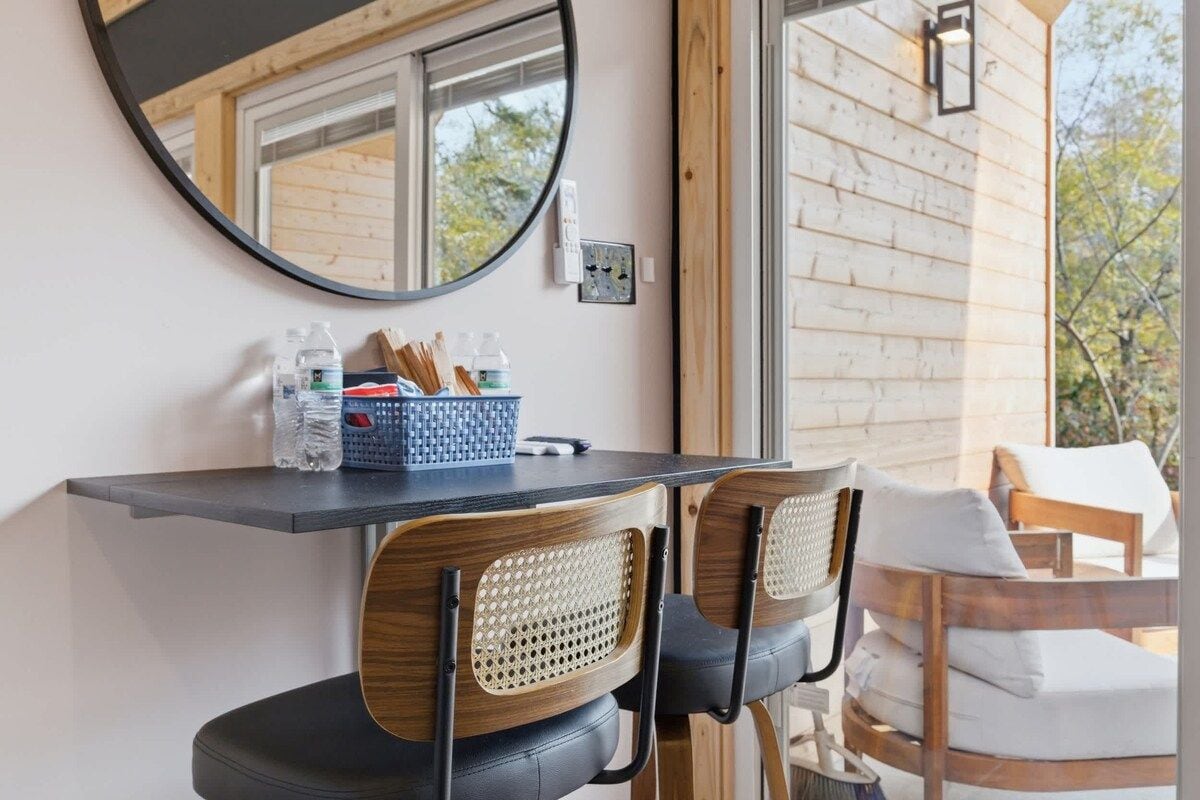 A room with a round mirror, a table with chairs, and a view of the outside.