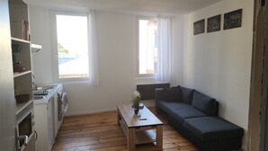 Ein kleiner, hell eingerichteter Raum mit Küchenzeile, einer Couch und einem Tisch mit Pflanze.