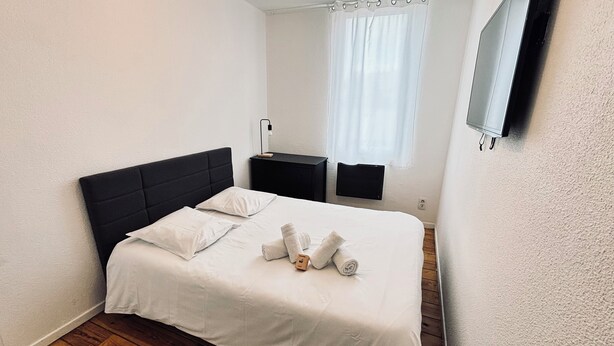 Ein ordentlich bezogenes Bett mit weißen Bettwäsche und Handtüchern, ein schwarzer Kopfteil, ein kleiner Nachttisch mit Lampe sowie ein Fenster mit weißem Vorhang.