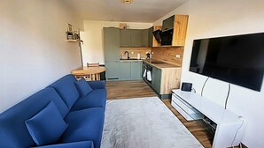 A compact living space with a blue sofa, a flat-screen TV, a kitchen area with wooden cabinets, and a dining table. - Charming apartment overlooking the valley (Briançon)