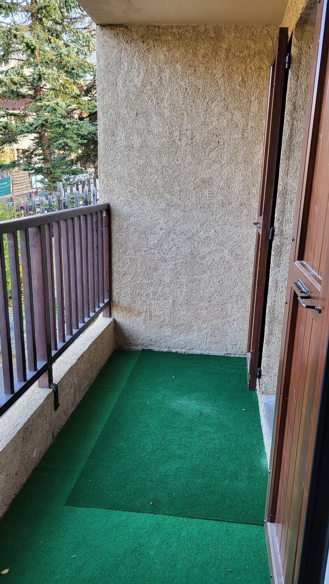 A small balcony with a green artificial turf floor, a wooden door, and a metal railing.