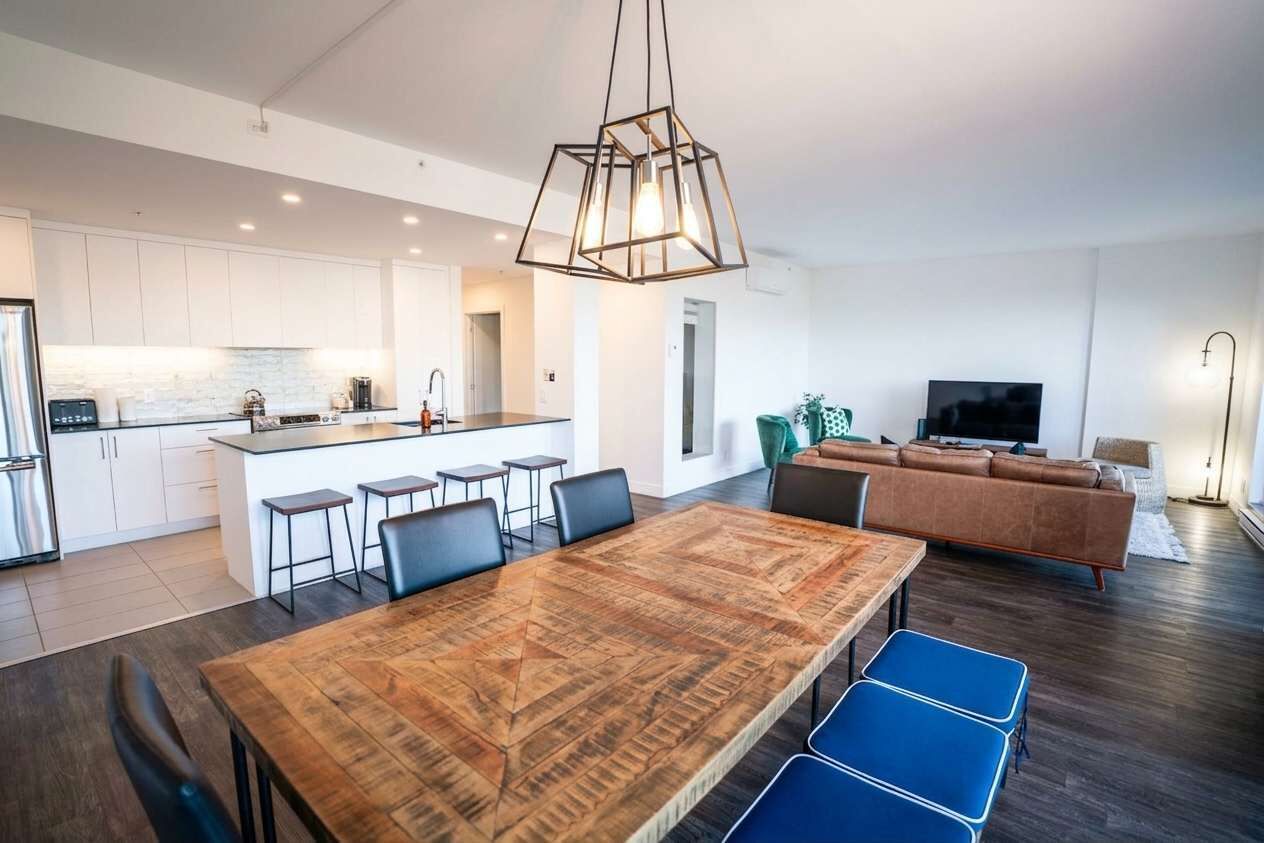 A modern kitchen with white cabinets, a central island with bar stools, a dining area with a wooden table and blue chairs, a living room with a brown sofa, and a TV.