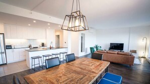 Eine moderne Küche mit weißen Schränken, einer zentralen Kochinsel mit Barhocker, einem Essbereich mit Holztisch und blauen Stühlen sowie einem Wohnzimmer mit braunem Sofa und Fernseher.