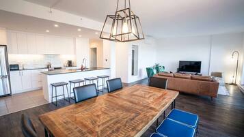 A modern kitchen with white cabinets, a central island with bar stools, a dining area with a wooden table and blue chairs, a living room with a brown sofa, and a TV.