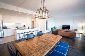 Eine moderne Küche mit weißen Schränken, einer zentralen Kochinsel mit Barhocker, einem Essbereich mit Holztisch und blauen Stühlen sowie einem Wohnzimmer mit braunem Sofa und Fernseher.