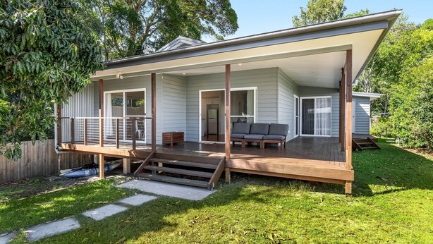 Ein modernes Haus mit überdachter Veranda, einer kleinen Terrasse und einer Glastür, die in einen Wohnbereich mit einer Couch führt.