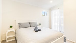Ein ordentlich gemachtes Bett mit gestreifter Bettdecke, ein kleiner Nachttisch mit einer Pflanze und ein Fenster mit durchscheinenden Vorhängen.