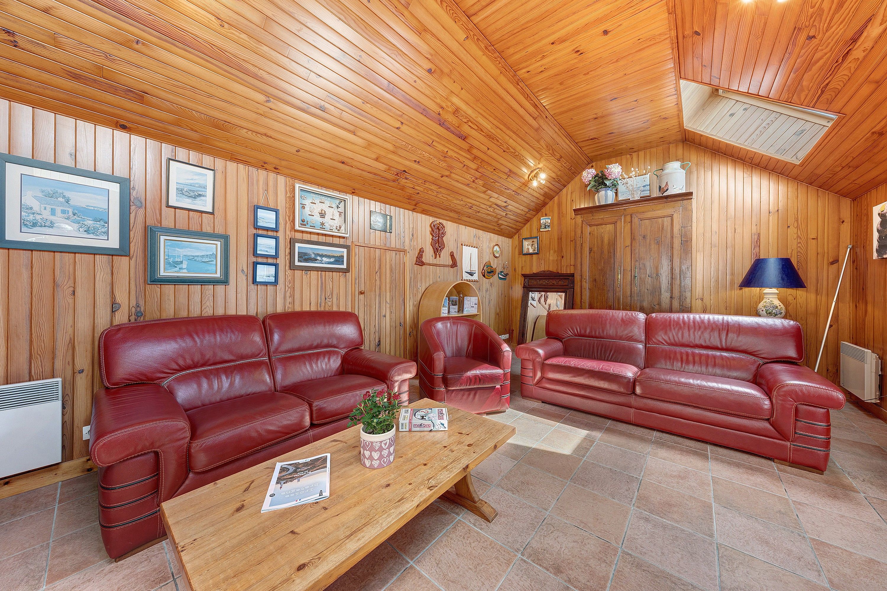 Une pièce chaleureuse aux murs et au plafond en bois, comprenant deux canapés en cuir rouge, une table basse en bois et divers tableaux encadrés.