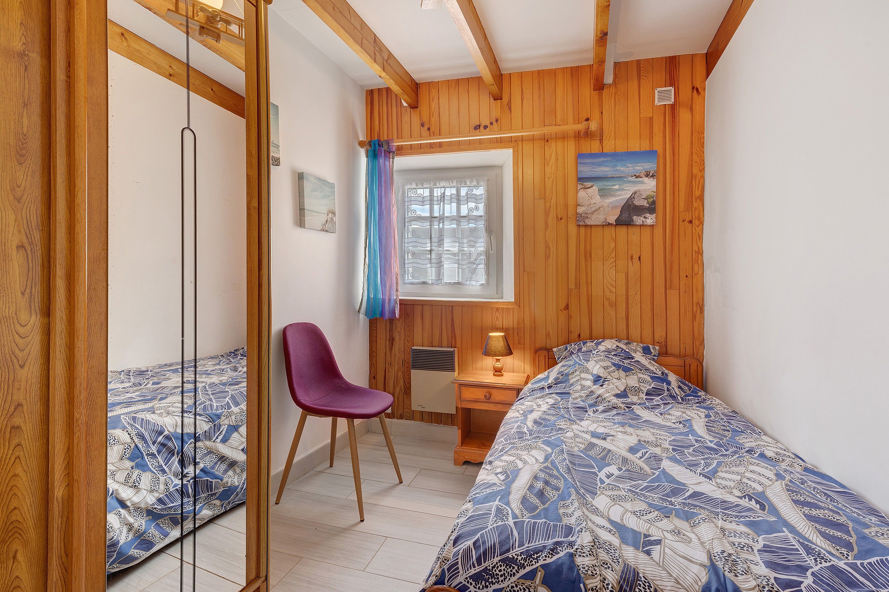 A bedroom with a wooden ceiling, a bed with a patterned bedspread, a chair, a desk with a lamp, and a window with a curtain.