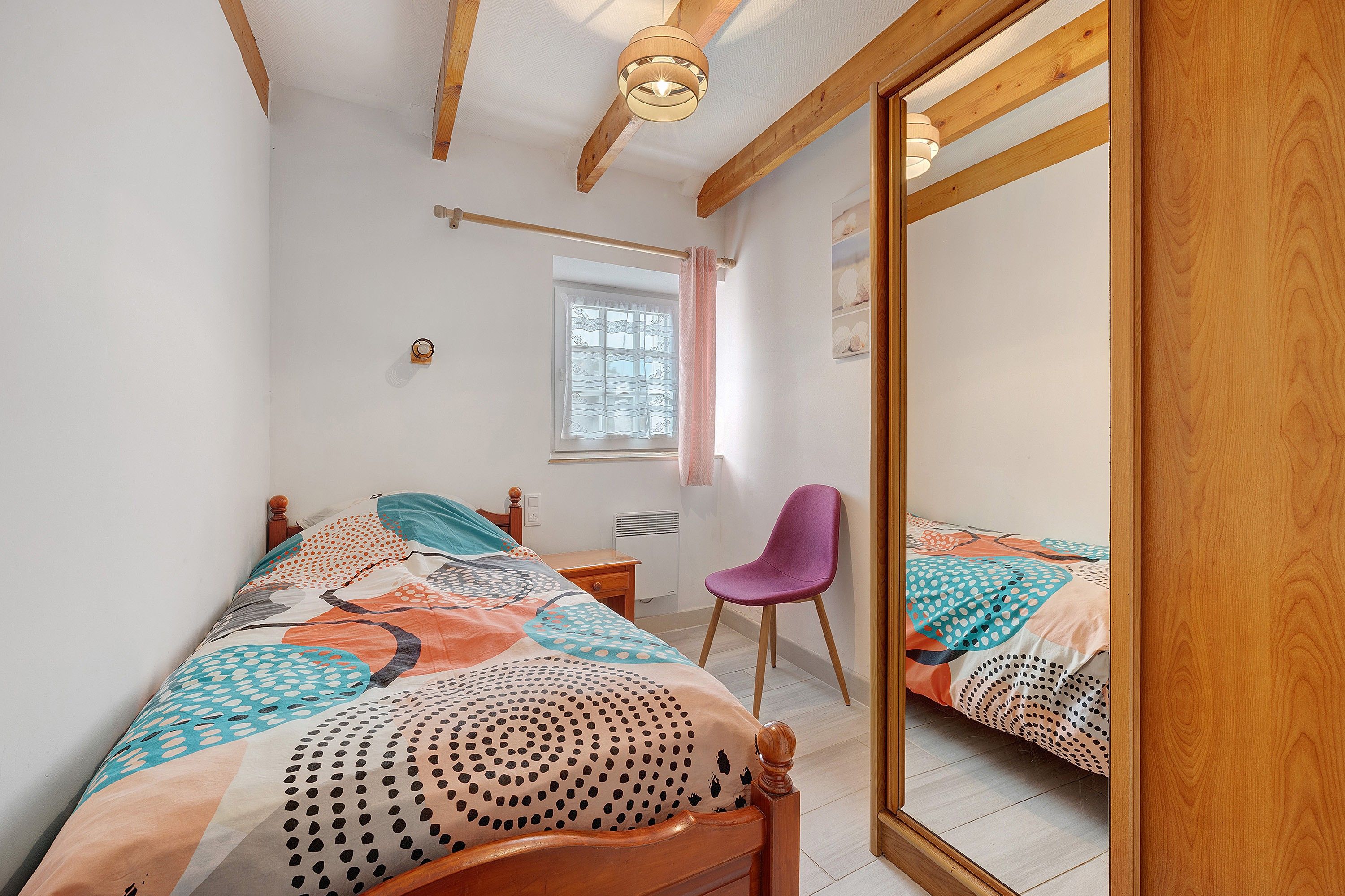 Een slaapkamer met een houten bed, een stoel, een raam met gordijnen en een spiegel.