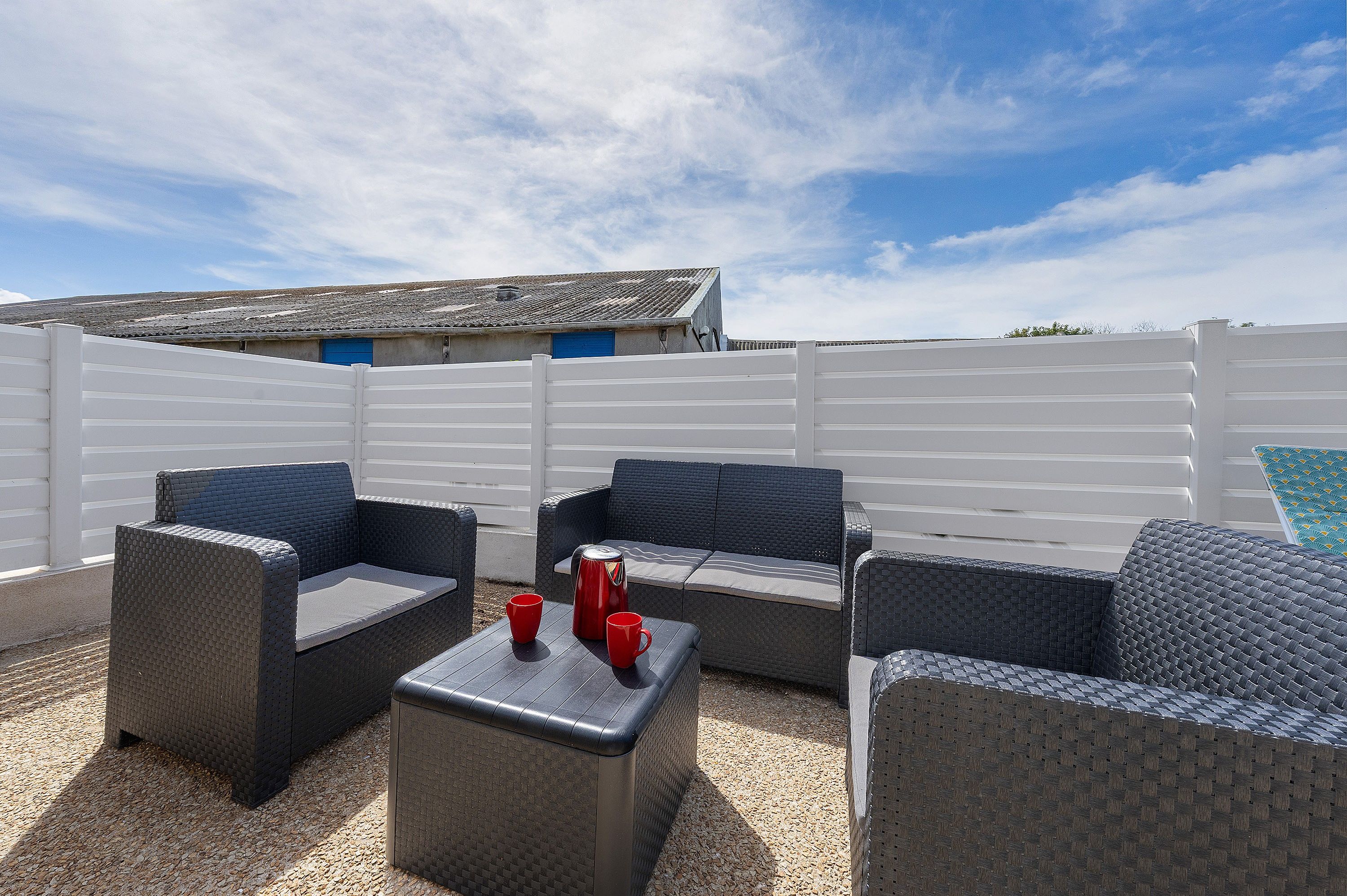 Een terras met rieten meubels en een bijzettafeltje, een wit hekwerk en een gebouw met een blauwe deur op de achtergrond.