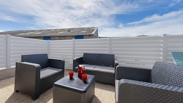 Een terras met rieten meubels en een bijzettafeltje, een wit hekwerk en een gebouw met een blauwe deur op de achtergrond.