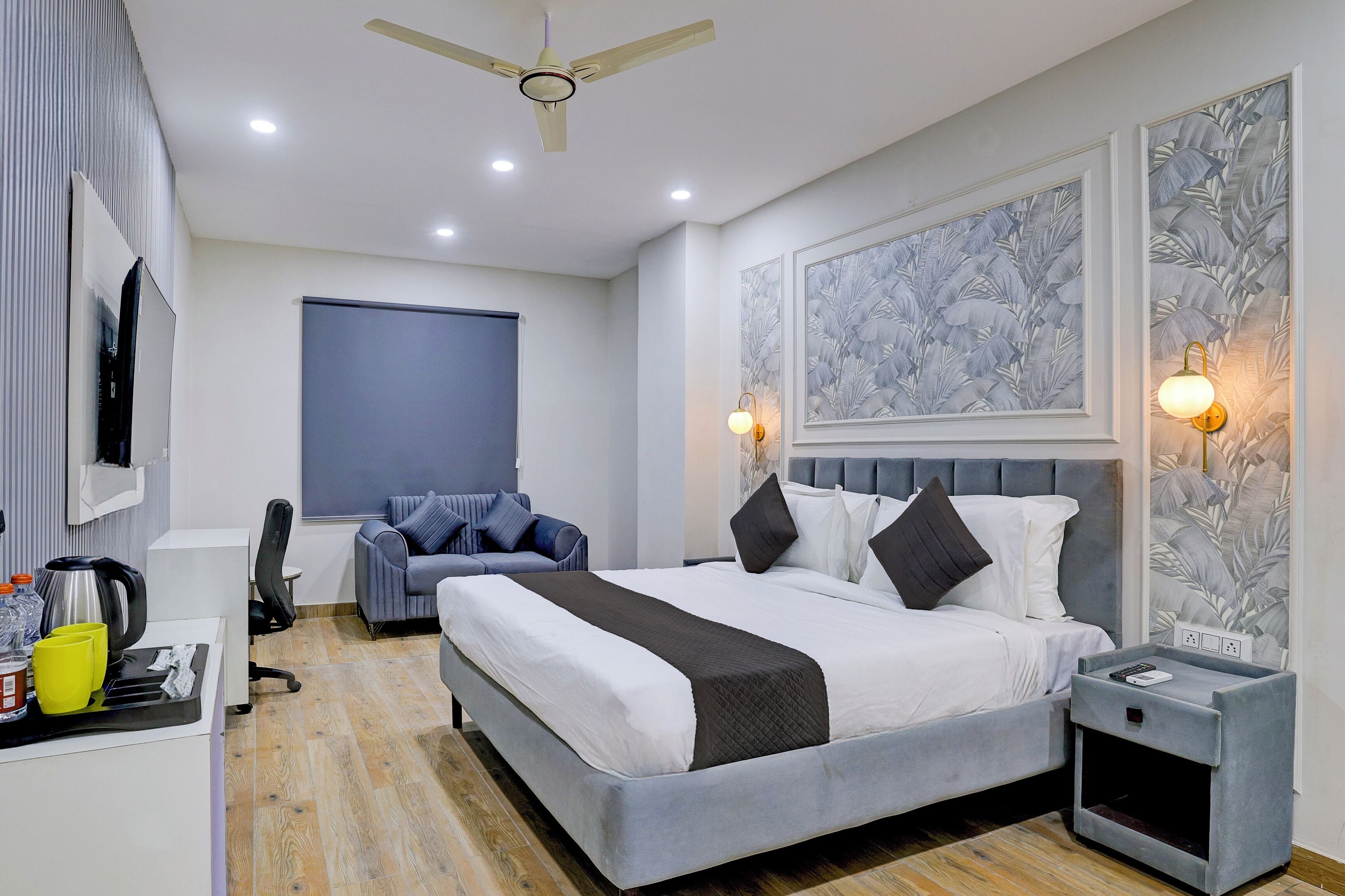 A modern hotel room with a large bed, a desk with a kettle, a chair, and a TV.