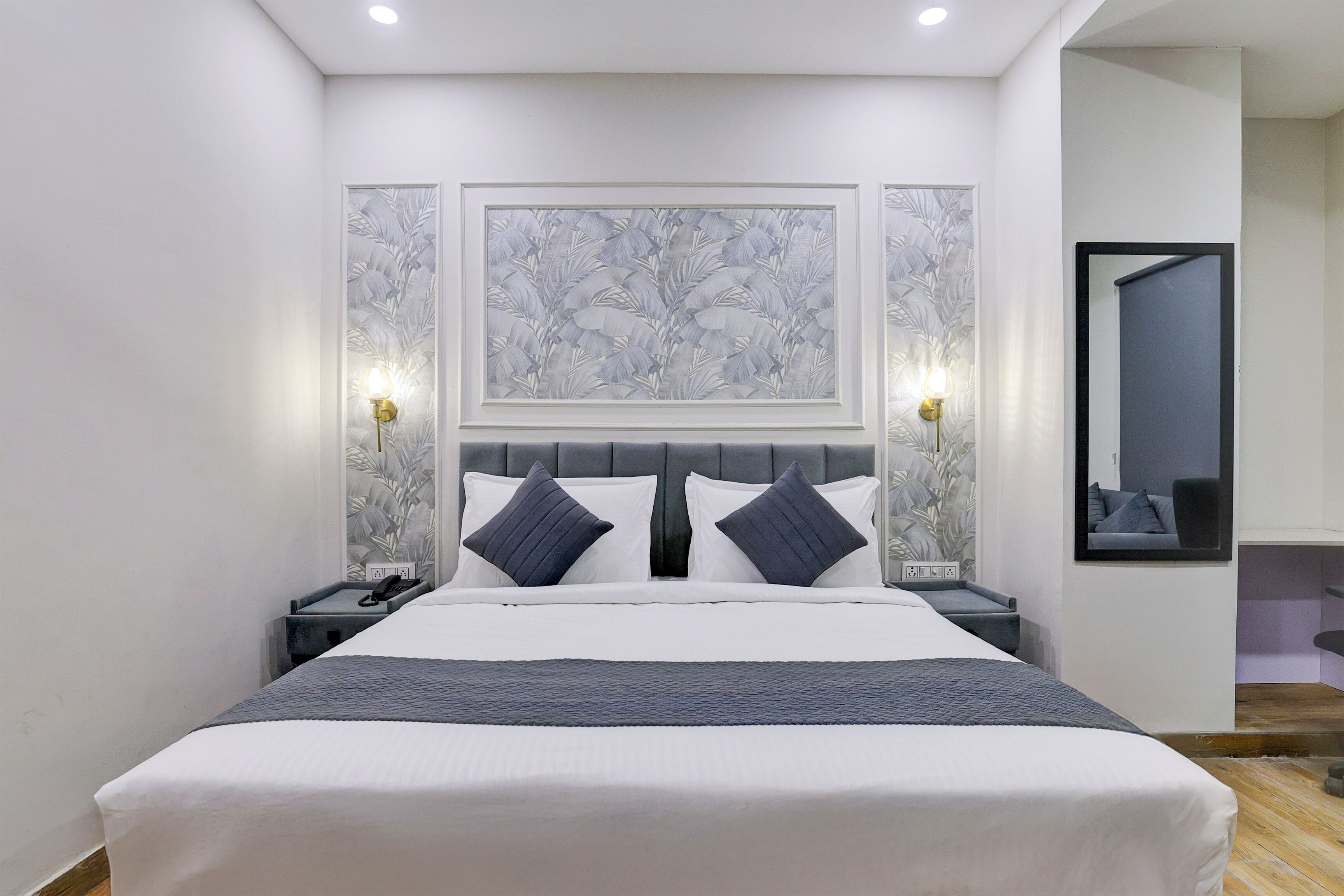 A neatly arranged bedroom with a large bed, two bedside tables, a nightstand, a wall-mounted mirror, and a framed floral artwork above the bed.