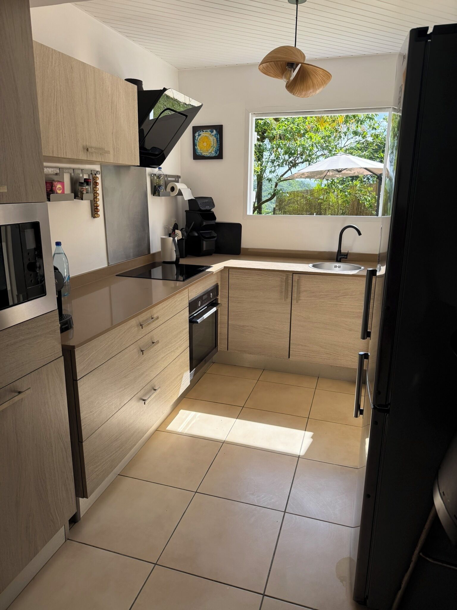 A modern kitchen with light wood cabinets, a black countertop, a built-in oven, and a window with a view of greenery.