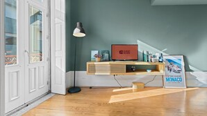 Ein Zimmer mit Holzboden, einer weißen Tür, einer grünen Wand, einer Stehlampe, einem Fernseher mit der Aufschrift „GUEST READY“ und einem gerahmten Bild von Monaco.