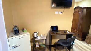 Una habitación compacta con kitchenette, un escritorio pequeño con una computadora portátil, una silla, un televisor montado en la pared y un armario de madera.