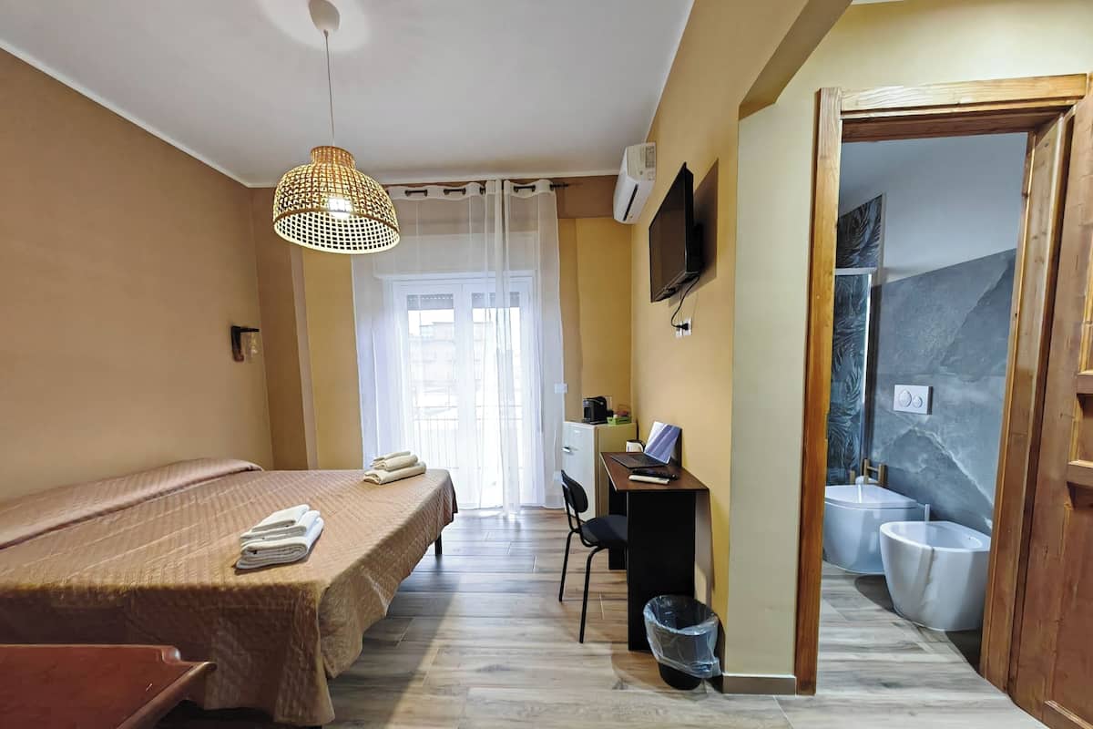 A hotel room with a bed, a desk with a computer, a TV, and a bathroom with a toilet and sink.