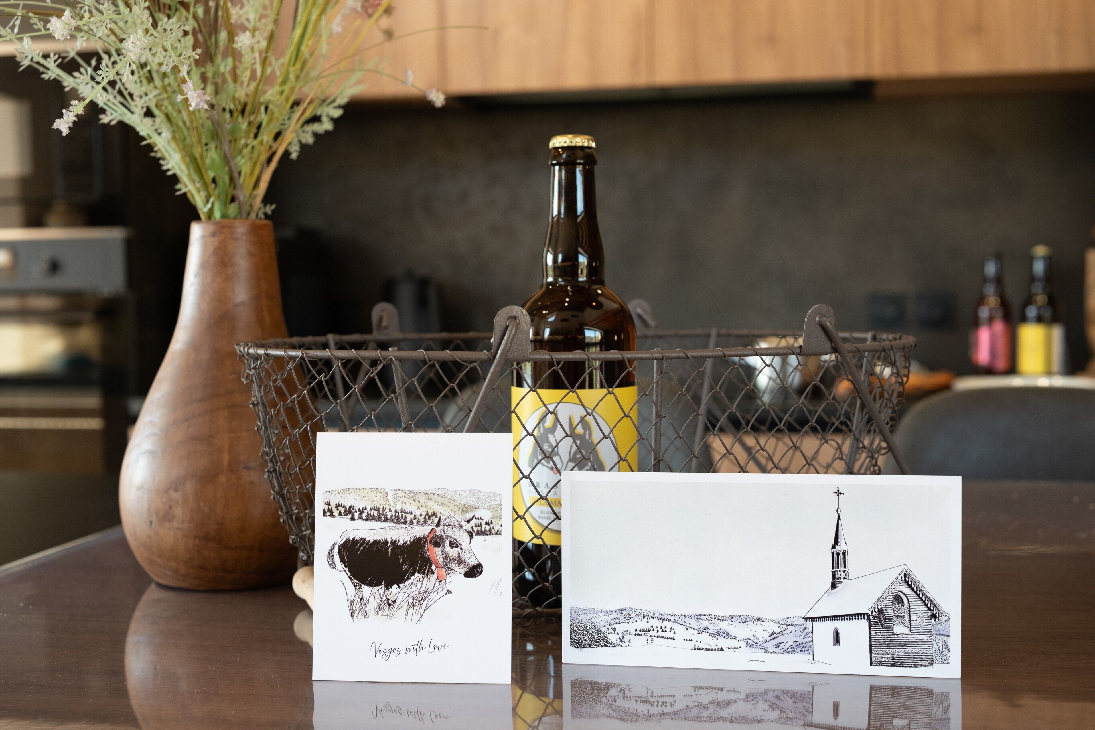A bottle of wine, two cards with illustrations, and a vase with dried flowers on a reflective surface.