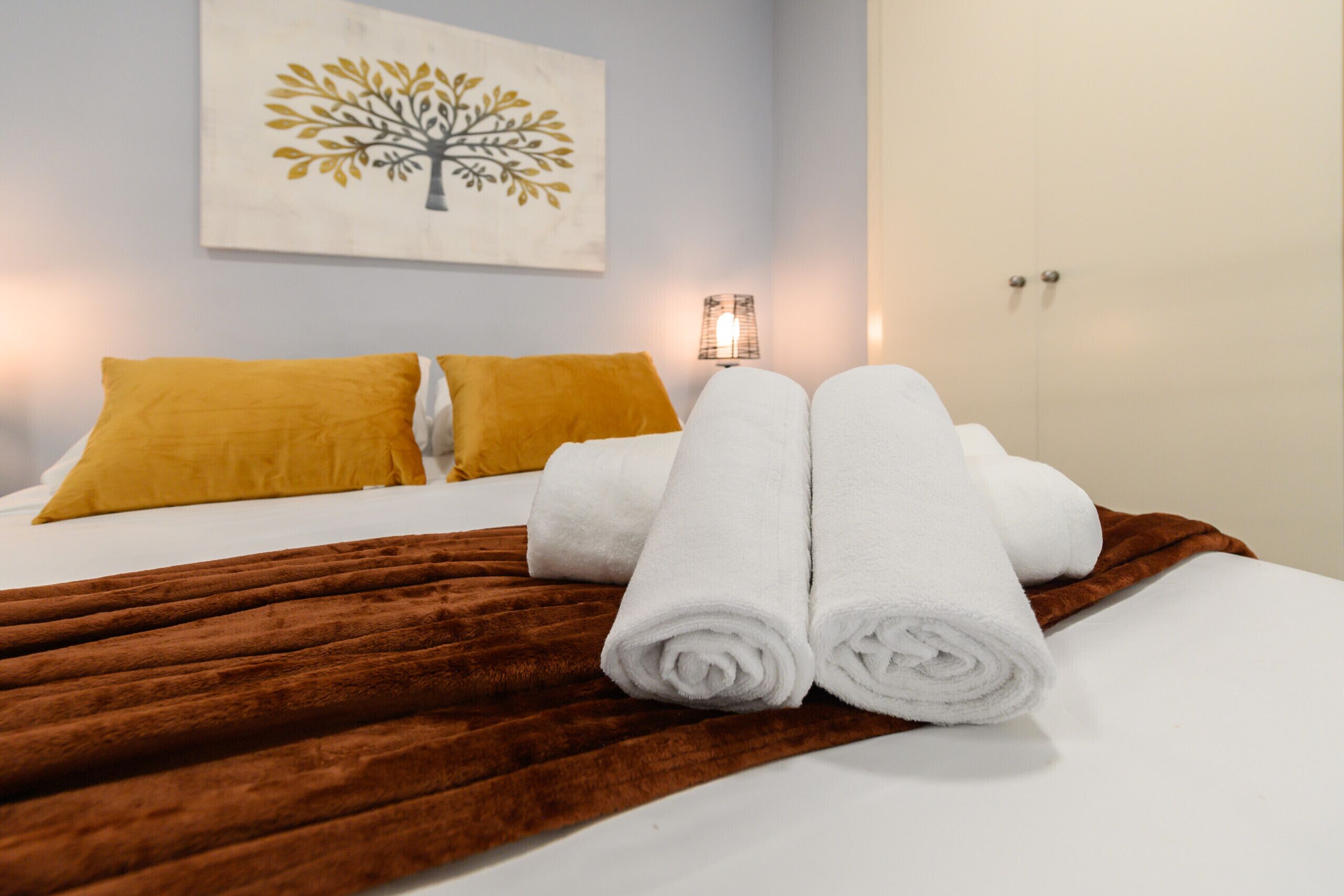 Een netjes opgemaakt bed met witte en mosterdkleurige kussens, een bruin plaid en opgerolde witte handdoeken.