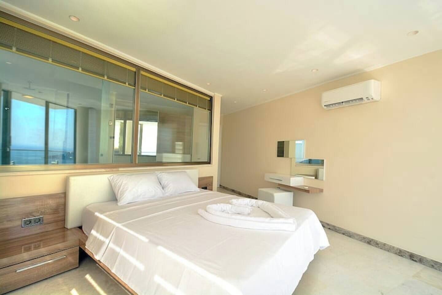 Ein modernes Hotelzimmer mit einem großen Bett, einem hölzernen Nachttisch, einer integrierten Sitzbank, einem großen Fenster und einer wandmontierten Klimaanlage.