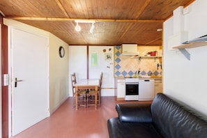 A room with a wooden ceiling, a dining table with chairs, a black leather sofa, a kitchen area with a stove and sink, and a wall clock.