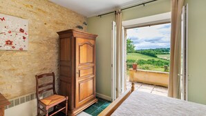 A bedroom with a large wooden wardrobe, a wooden chair, a bed, and a view of a green landscape through an open door. - Gite Charly: Private Terrace, Shared Garden, Pool & Wi-Fi in Salignac Eyvigues (Salignac Eyvigues)