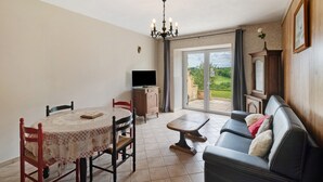 A dining area with a table and chairs, a sofa, a television, and a view through a glass door to the outside. - Gîte Charly: Private Terrace, Shared Garden, Pool & Wi-Fi in Salignac Eyvigues (Salignac Eyvigues)