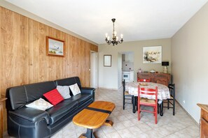 Un salon avec un canapé en cuir noir, une table basse en bois, une table à manger avec des chaises, un lustre et, au fond, un coin cuisine.