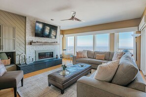 Un salón espacioso con chimenea, un televisor de pantalla plana grande montado en una pared texturizada, un conjunto de sofás cómodos y vistas de la ciudad a través de amplios ventanales.