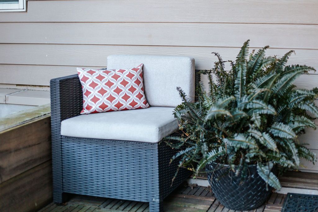 A wicker chair with a cushion and a patterned pillow, a potted fern plant, and a wooden deck.