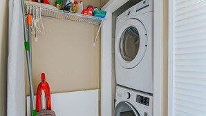 Een wasruimte met een stapel wasmachine en droger, een dweil, een bezem en een plank met schoonmaakmiddelen.